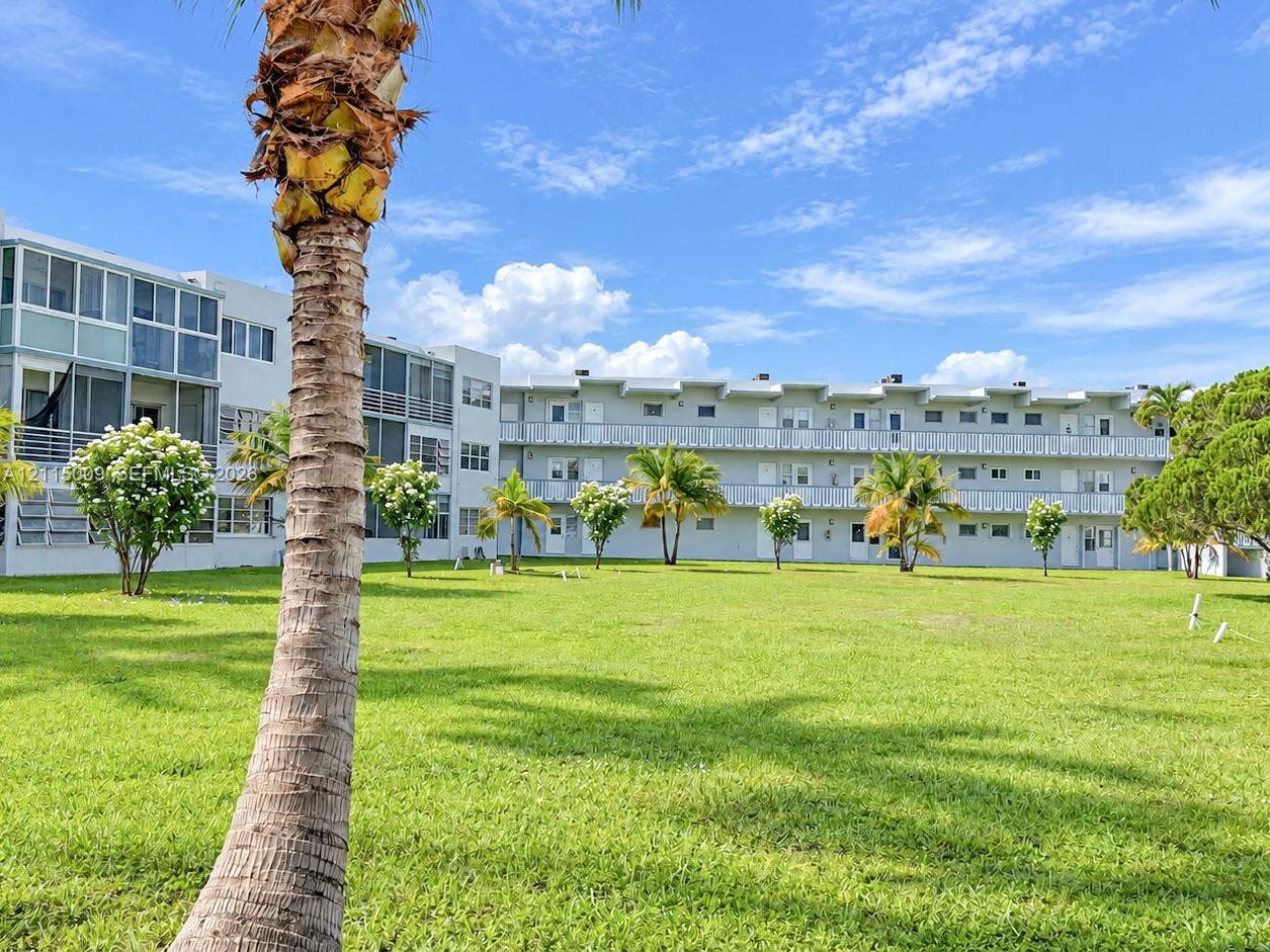 Multi-level exterior featuring light-colored facade, screened balcony enclosures, exterior metal railings, and expansive common grass area with mature palm trees