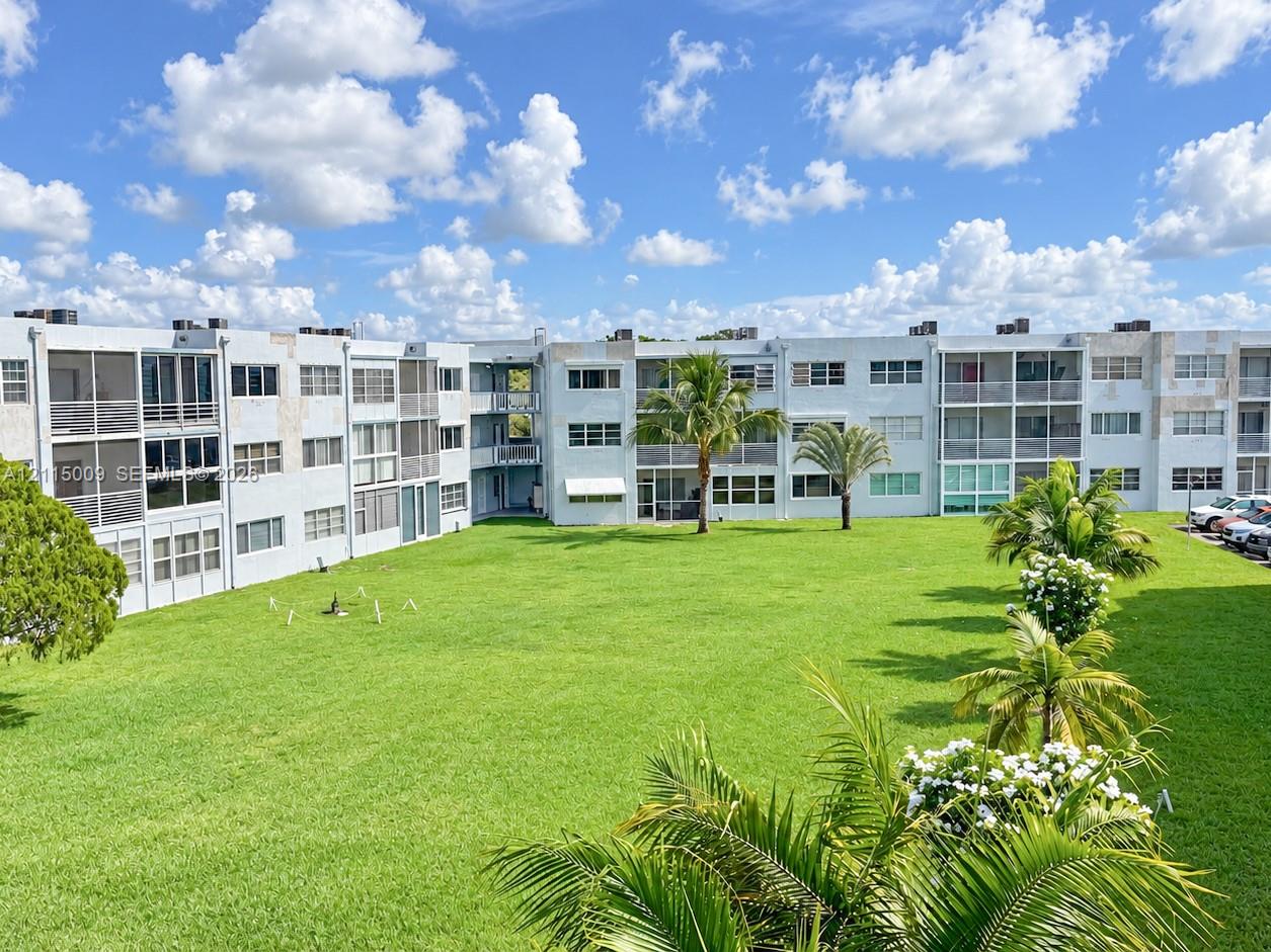 Multi-story residential building featuring screened balconies, expansive communal lawn, mature palm trees, and structural stucco exterior