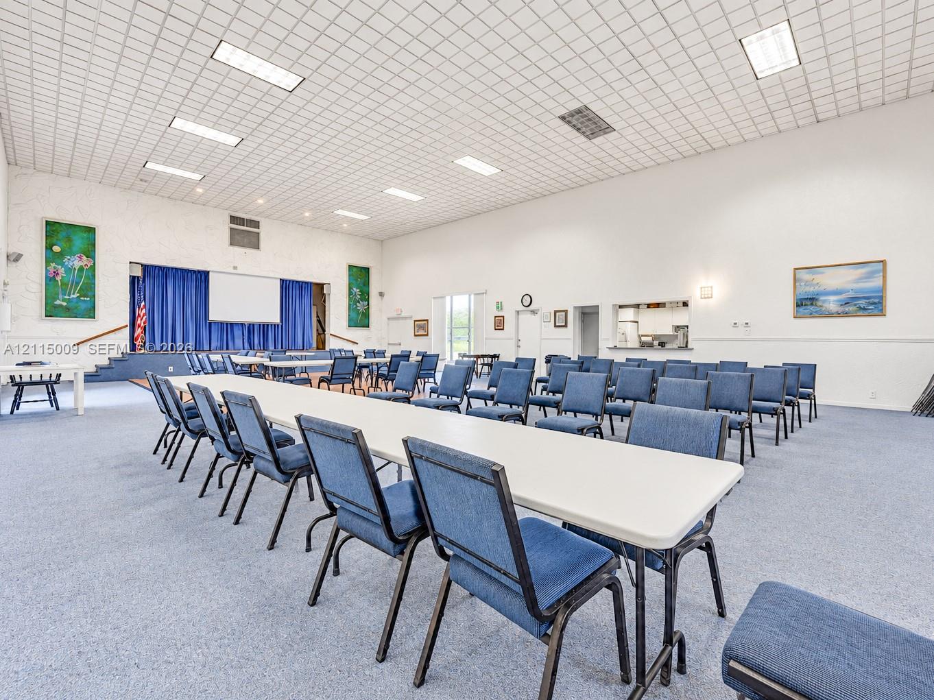 Expansive multi-purpose hall featuring a raised stage with curtain backdrop, recessed grid ceiling lighting, carpeted flooring, and a service pass-through window