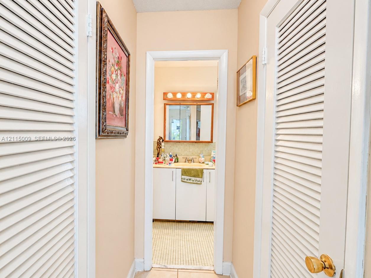 Hallway featuring dual white louvered closet doors and tile flooring, opening to a vanity area with white cabinetry, a framed vanity mirror, and a wall-mounted light fixture