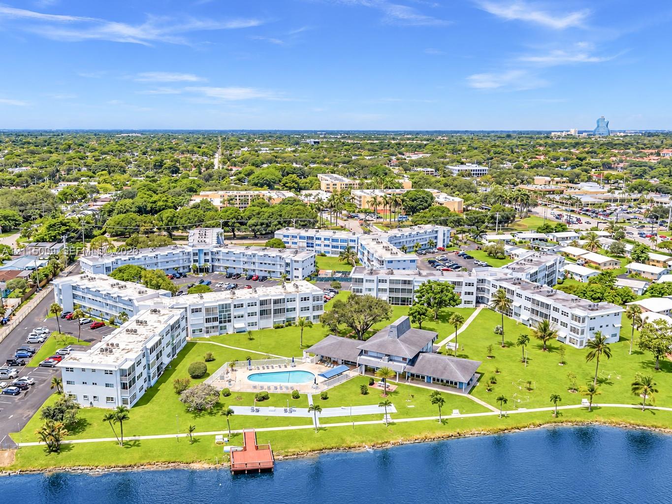 Aerial perspective of a multi-building residential complex featuring a community swimming pool, waterfront dock, expansive green space, and paved surface parking