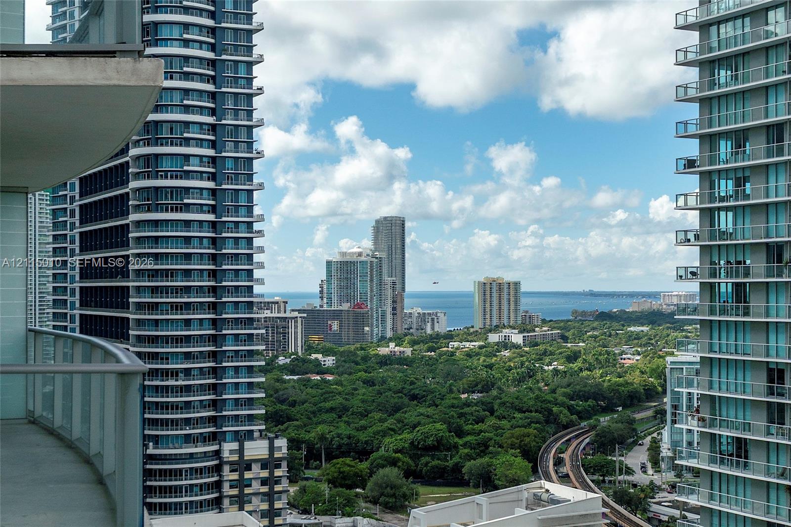 Private balcony featuring concrete flooring and a glass-paneled railing overlooking an urban skyline, lush forest canopy, and coastal water