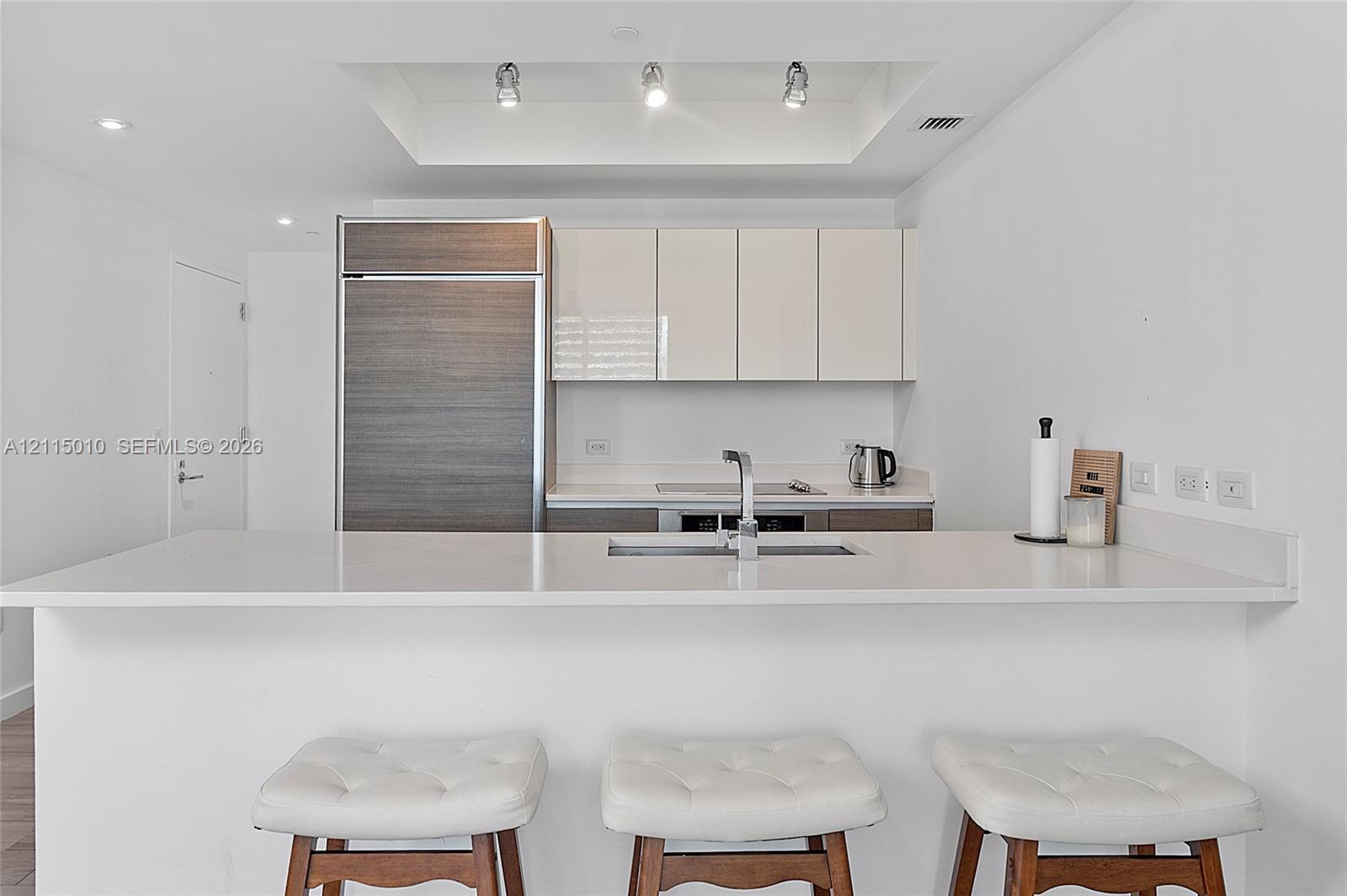 Kitchen featuring a solid-surface countertop, integrated refrigerator, flat-panel cabinetry, recessed tray ceiling lighting, and a dual-basin undermount sink