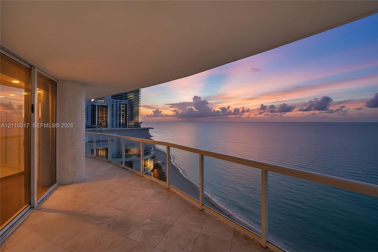 Curved private balcony featuring stone flooring, a glass railing, and expansive ocean and coastal scenery