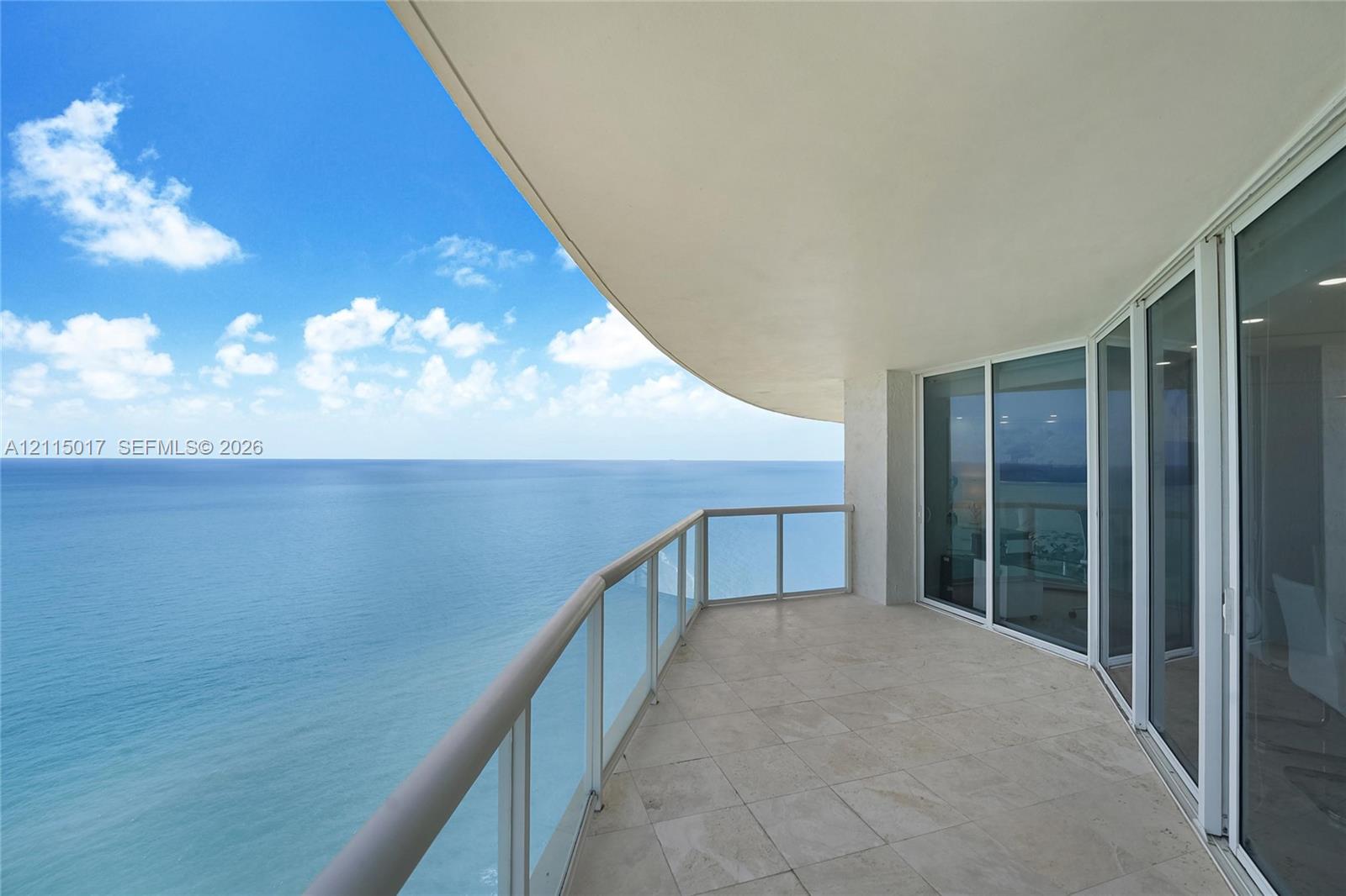 Curved terrace featuring stone tile flooring, floor-to-ceiling glass sliding doors, and a glass-paneled railing overlooking the ocean