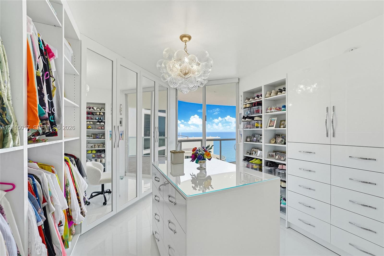 Custom dressing room featuring floor-to-ceiling white cabinetry, mirrored wardrobe doors, a central storage island with a glass top, and a multi-globe glass chandelier