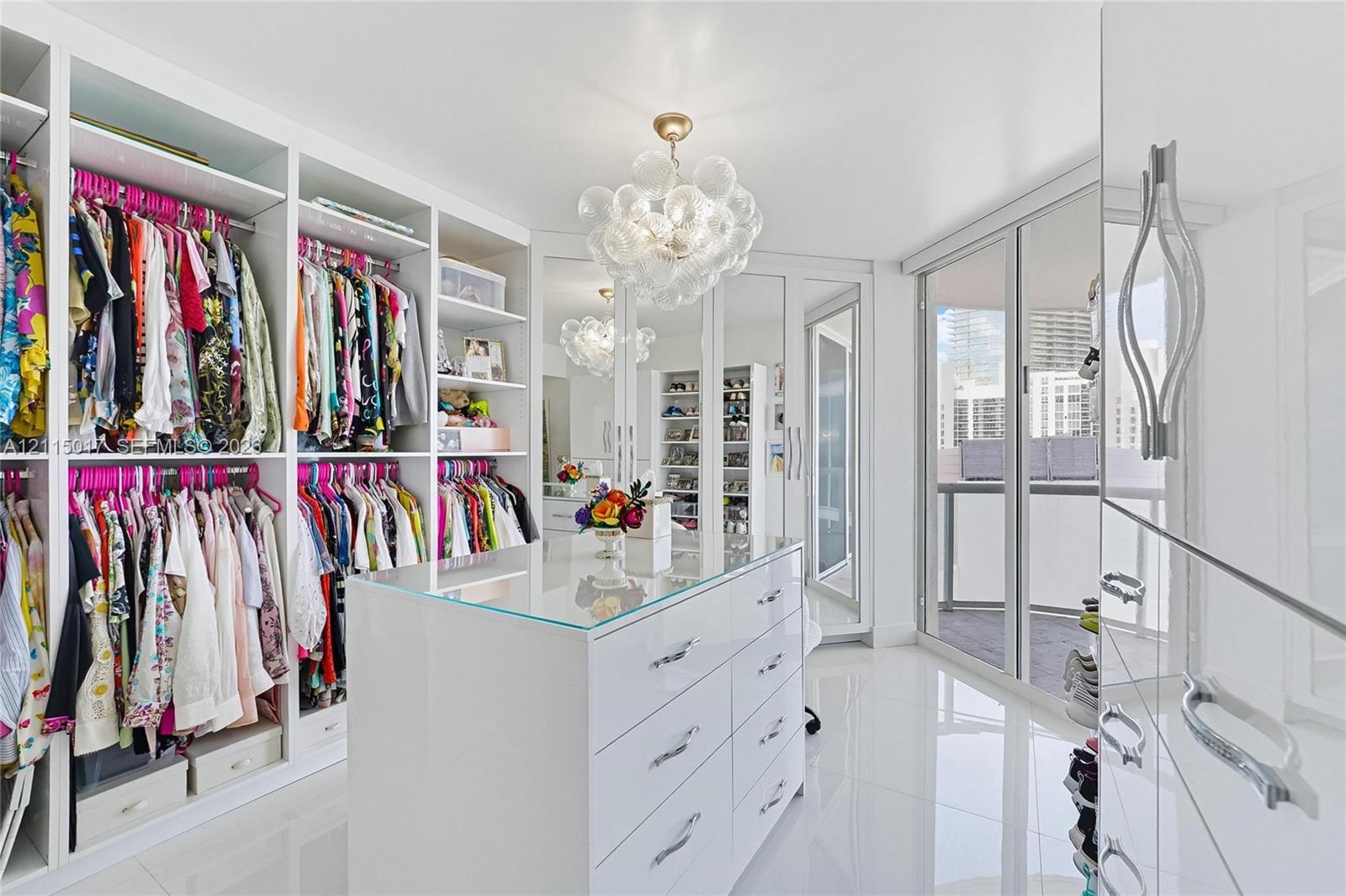 Custom built-in storage system with floor-to-ceiling shelving, central island featuring a glass top and drawers, mirrored cabinet doors, modern glass bubble chandelier, and large-format tile flooring