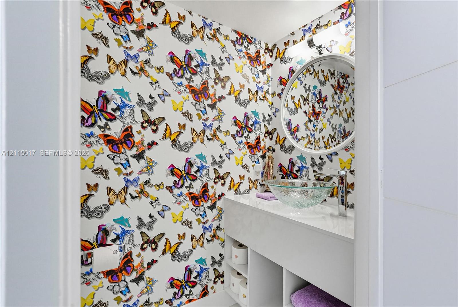 Powder room featuring butterfly-patterned wallpaper, a glass vessel sink, a quartz countertop, open built-in lower cabinetry, and a circular wall-mounted mirror