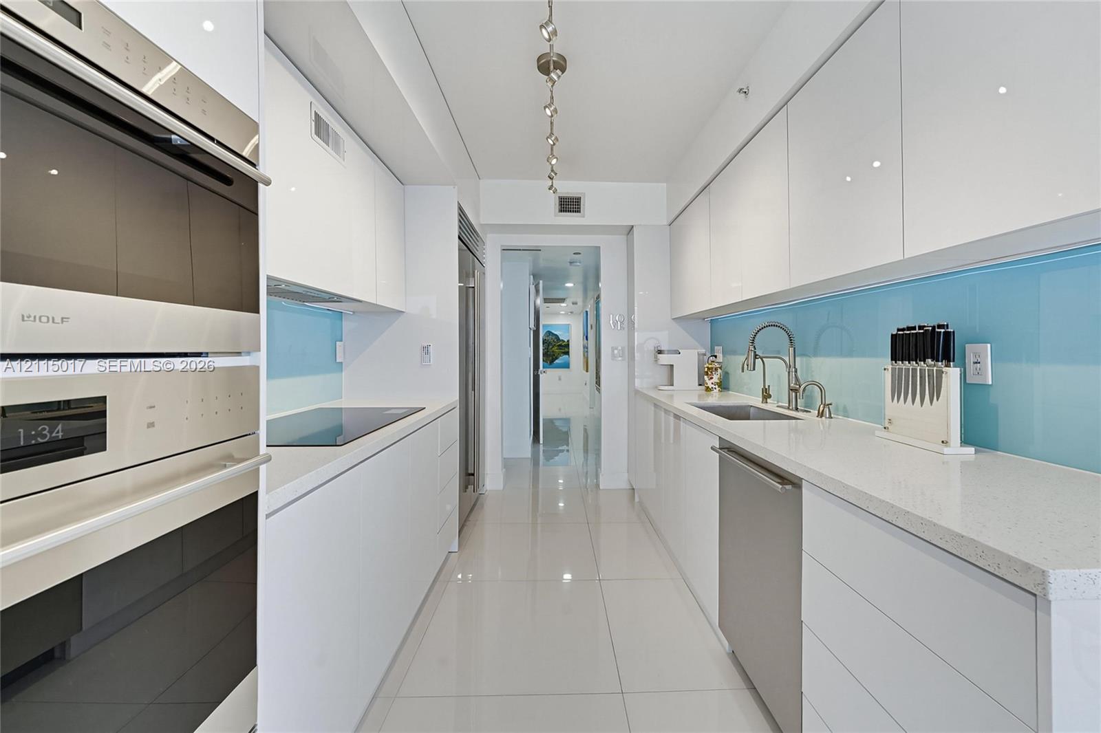 Galley kitchen featuring high-gloss white cabinetry, polished light-tone tile flooring, solid-surface countertops, blue glass backsplash, and integrated professional-grade appliances