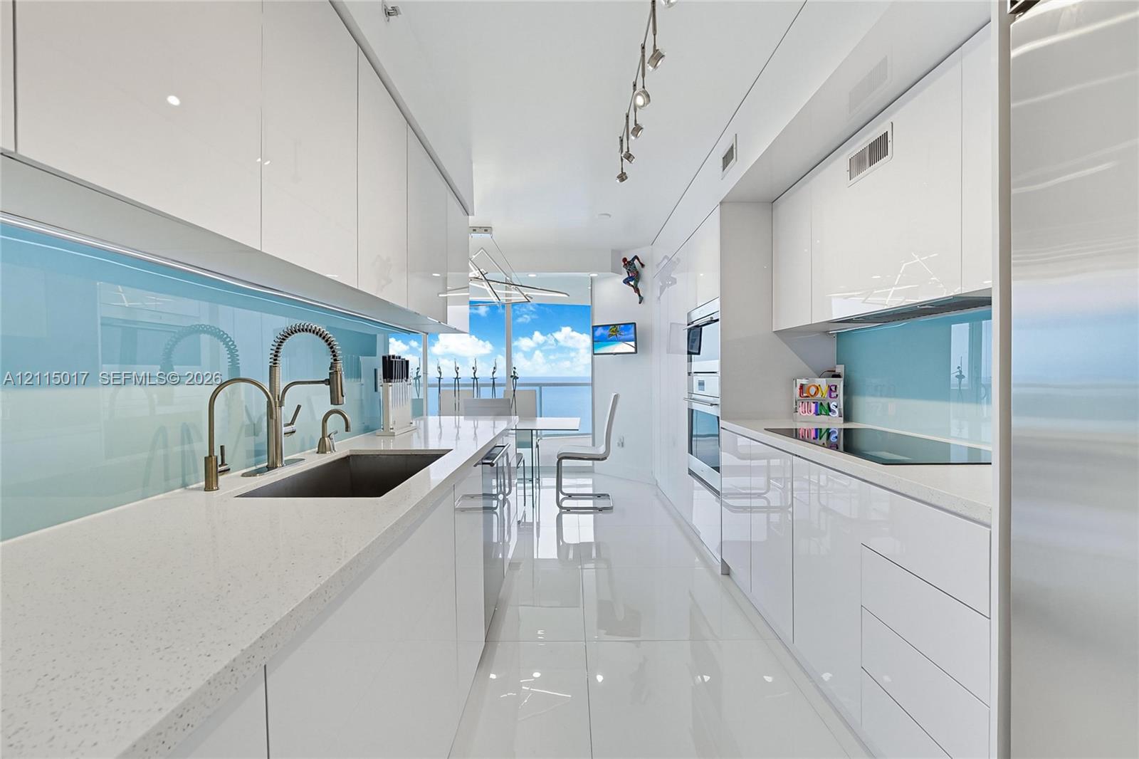 Galley kitchen featuring high-gloss white cabinetry, white stone-composite countertops, blue glass backsplash, stainless steel undermount sink with gooseneck faucet, and built-in stainless steel double wall ovens