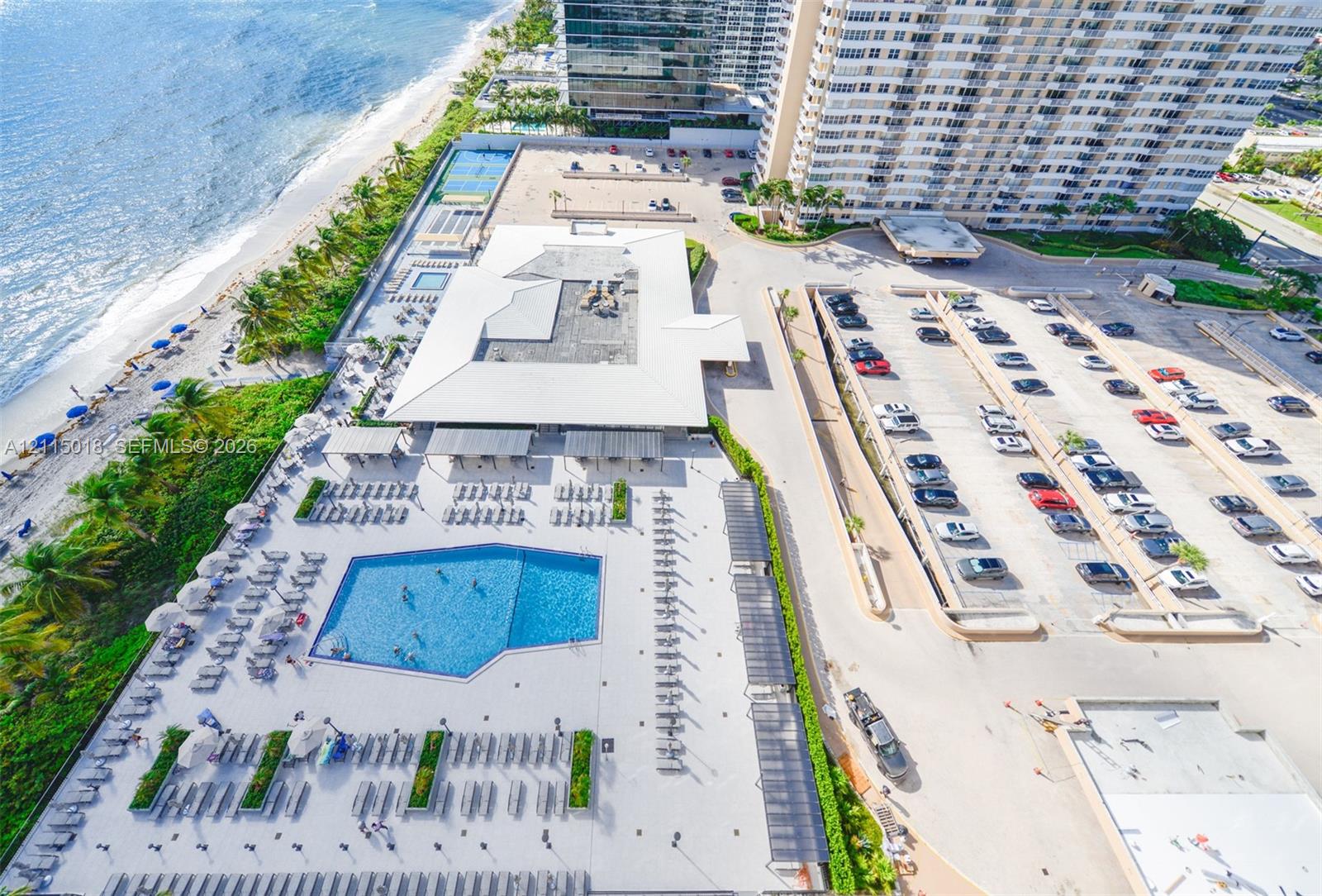 Aerial perspective of coastal property grounds featuring a swimming pool, paved sundeck, dedicated surface parking, and tennis courts