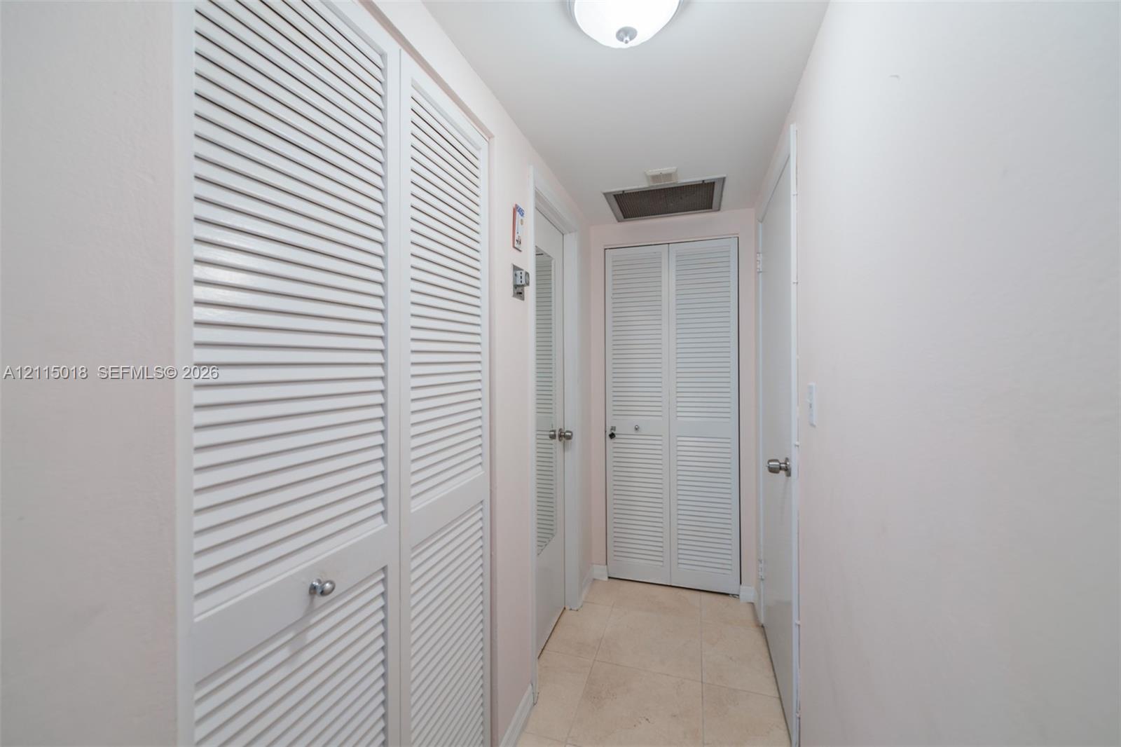Hallway featuring neutral tile flooring, multiple louvered-style bi-fold closet doors, and a flush-mount ceiling light fixture