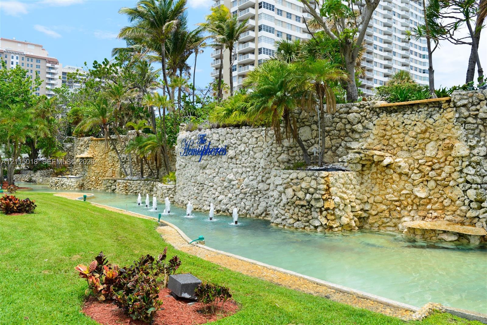Landscaped grounds featuring a tiered stone retaining wall, water feature with integrated fountains, manicured lawn, and mature palm trees