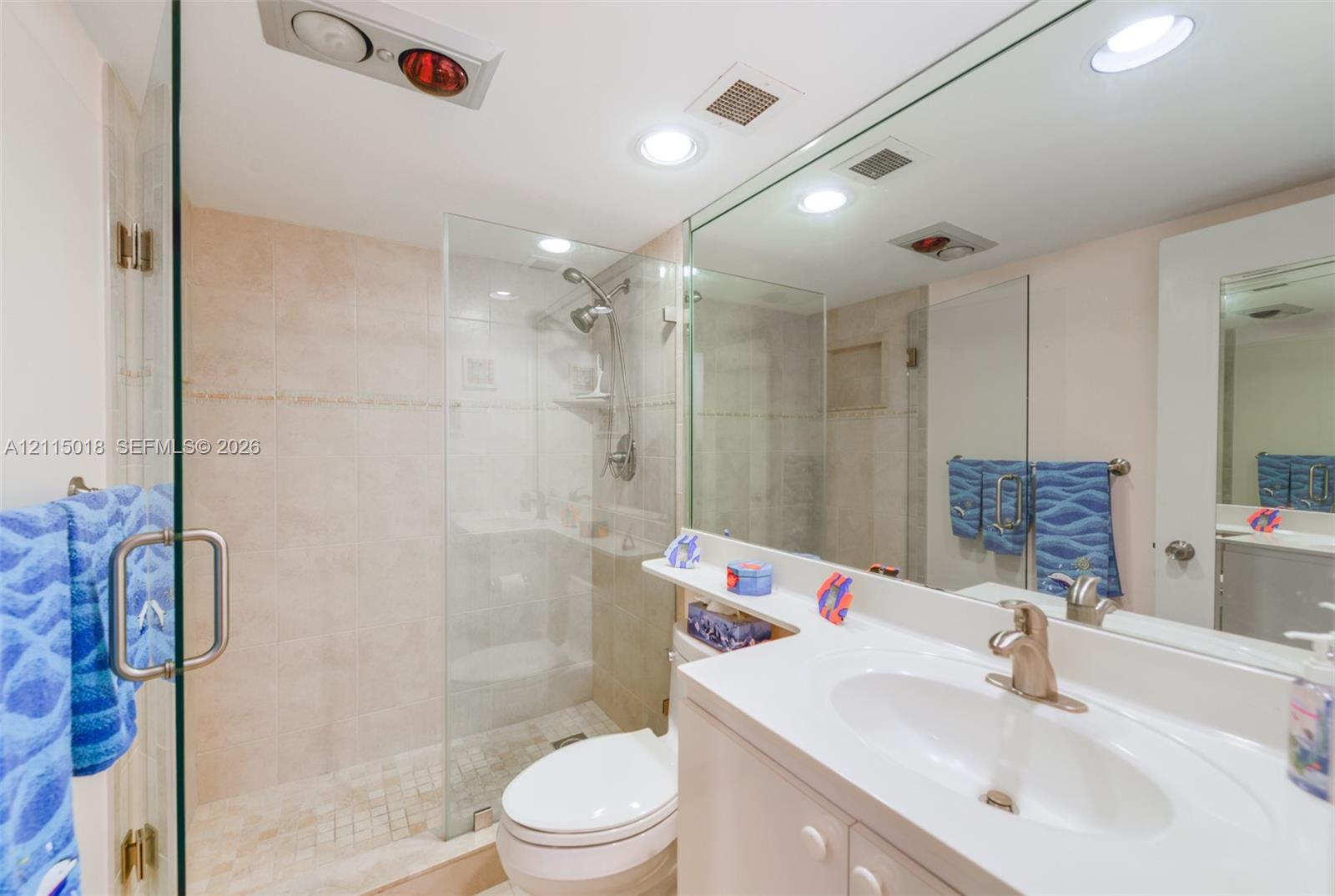 Full-width mirror over a white vanity with an integrated basin, glass-enclosed shower featuring stone-finish wall tile, and recessed lighting