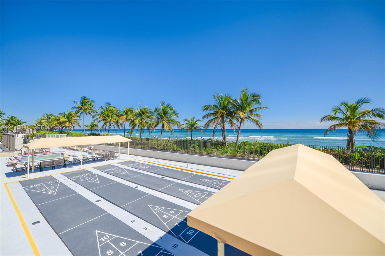 Outdoor recreation area featuring two paved shuffleboard courts, covered seating structures, and oceanfront placement