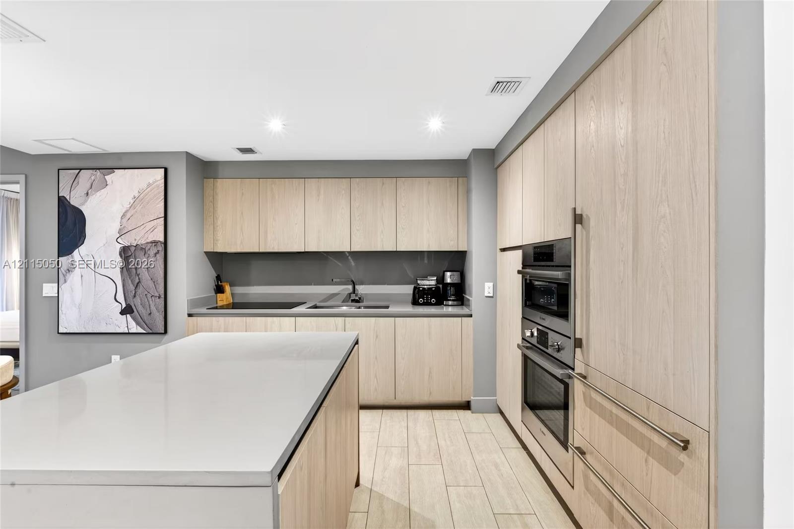 Kitchen featuring light-toned wood cabinetry, stone-surface island, built-in double wall oven, cooktop, and wood-finish flooring Kitchen featuring light-toned wood cabinetry, stone-surface island, built-in double wall oven, cooktop, and wood-finish flooring