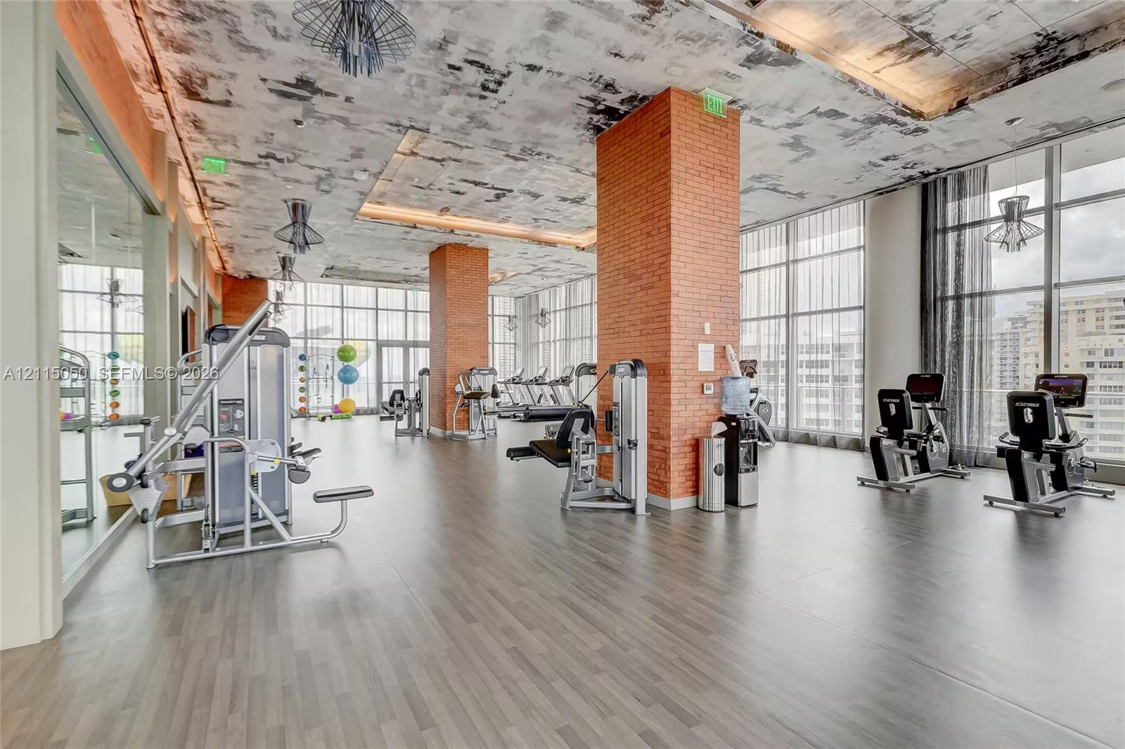 Fitness facility featuring floor-to-ceiling windows, exposed brick structural pillars, industrial-style concrete textured ceiling, and wood-finish flooring