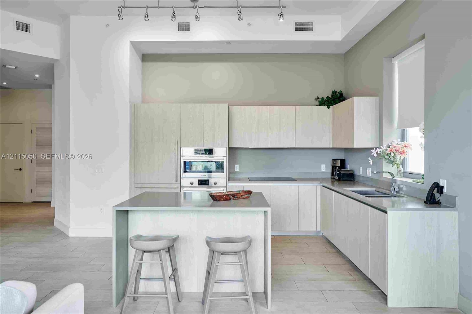 Kitchen featuring light-finish cabinetry, a waterfall-edge island, integrated stainless steel appliances, track lighting, and tile flooring Kitchen featuring light-finish cabinetry, a waterfall-edge island, integrated stainless steel appliances, track lighting, and tile flooring