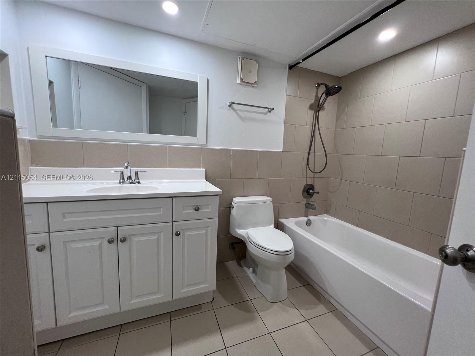 Bathroom featuring a white vanity with integrated basin, large rectangular wall mirror, tile-surround bathtub with a detachable handheld showerhead, and neutral tile flooring