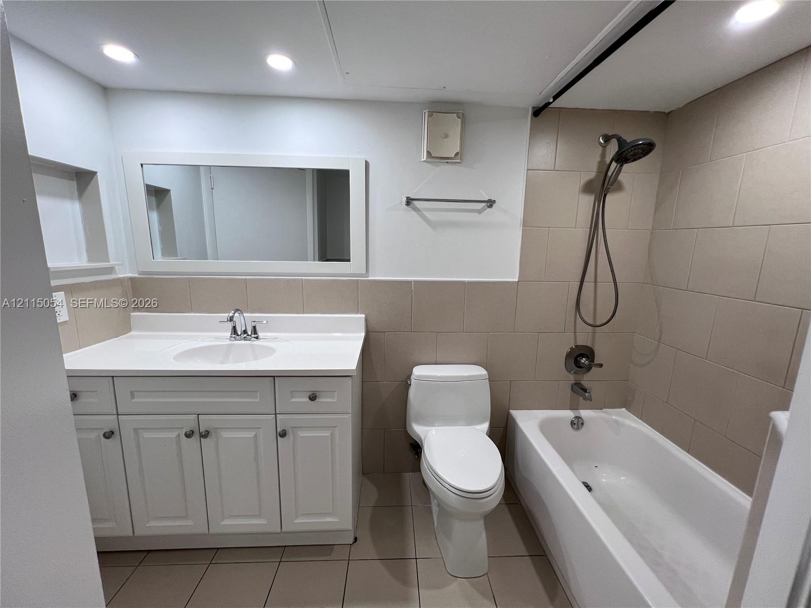 Bathroom featuring a white vanity cabinet with an integrated countertop sink, wall-mounted rectangular mirror, recessed lighting, and a neutral tile surround in the shower area