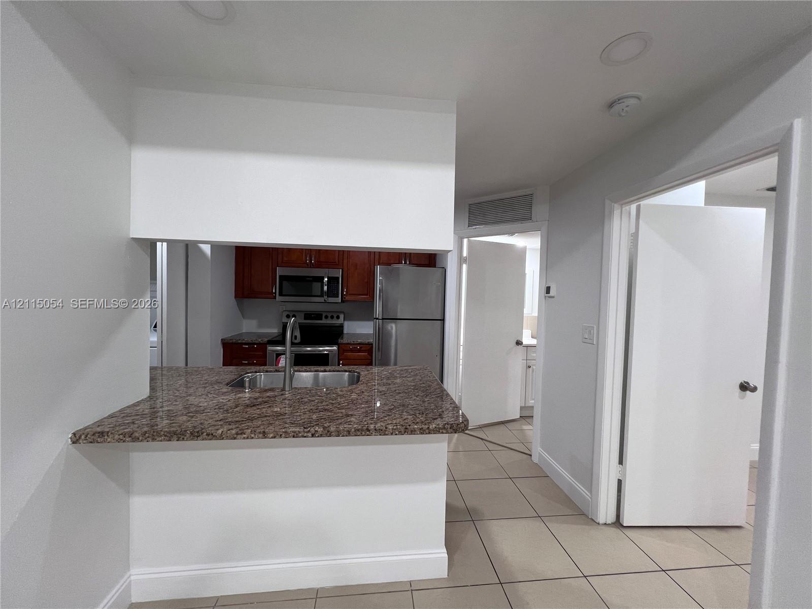 Pass-through kitchen counter with a stone-finish surface, stainless steel double-basin sink, wood-finish cabinetry, stainless steel appliances, and ceramic tile flooring