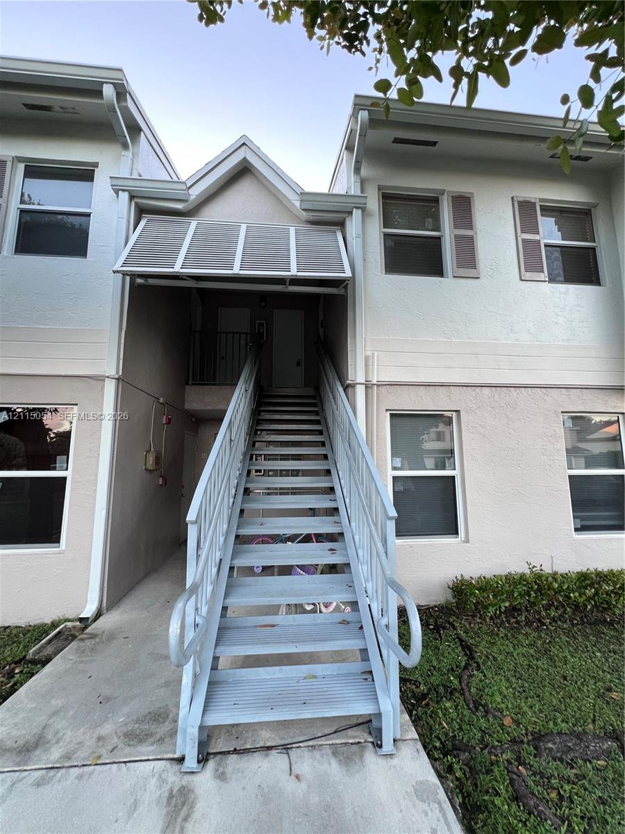 Two-story exterior featuring light-toned stucco siding, metal staircase with powder-coated railings, integrated window shutters, and a fixed metal awning over the entryway