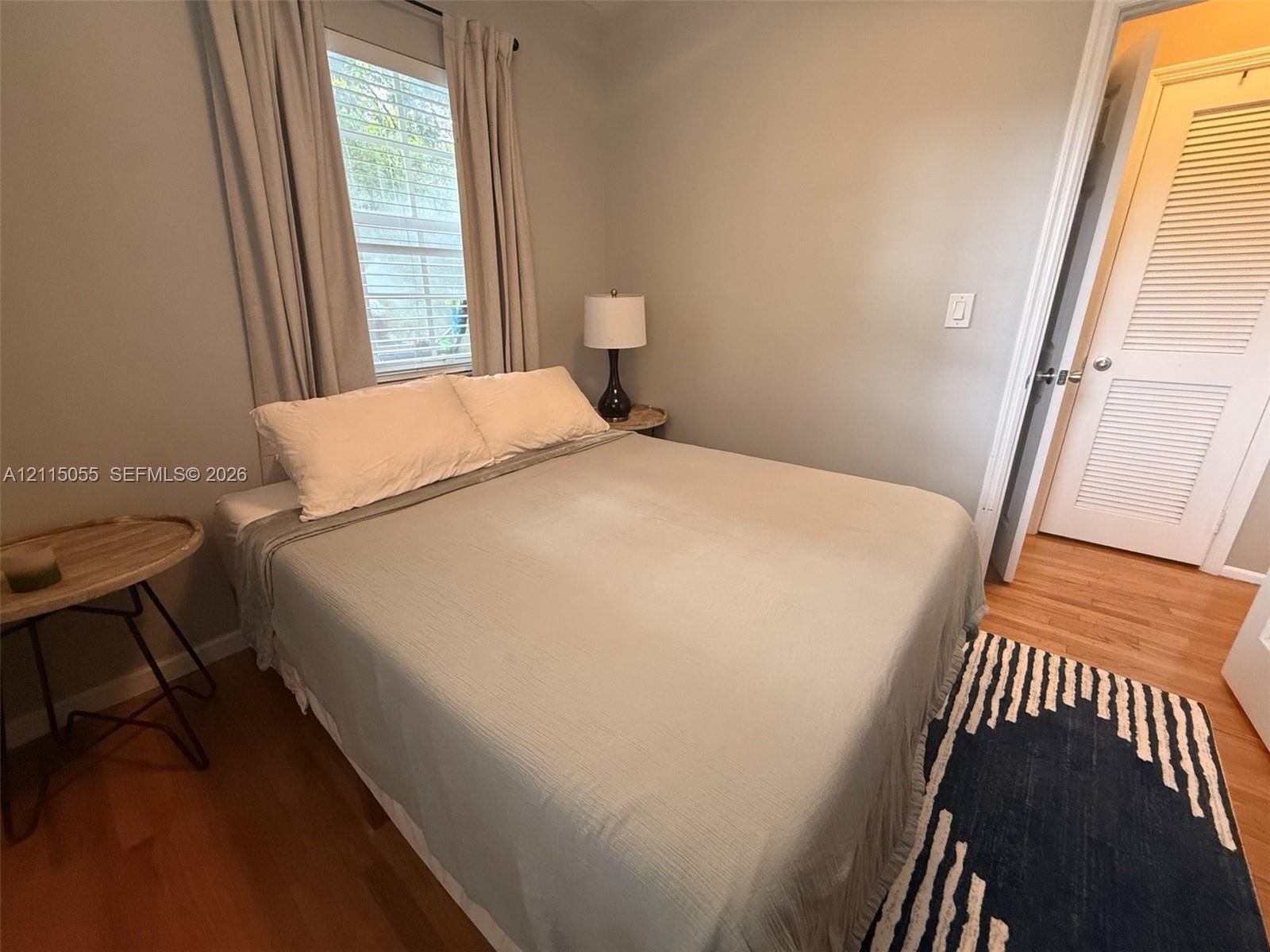 Neutral-toned interior room featuring wood-finish flooring, a single window with horizontal blinds, and a louvered-panel door