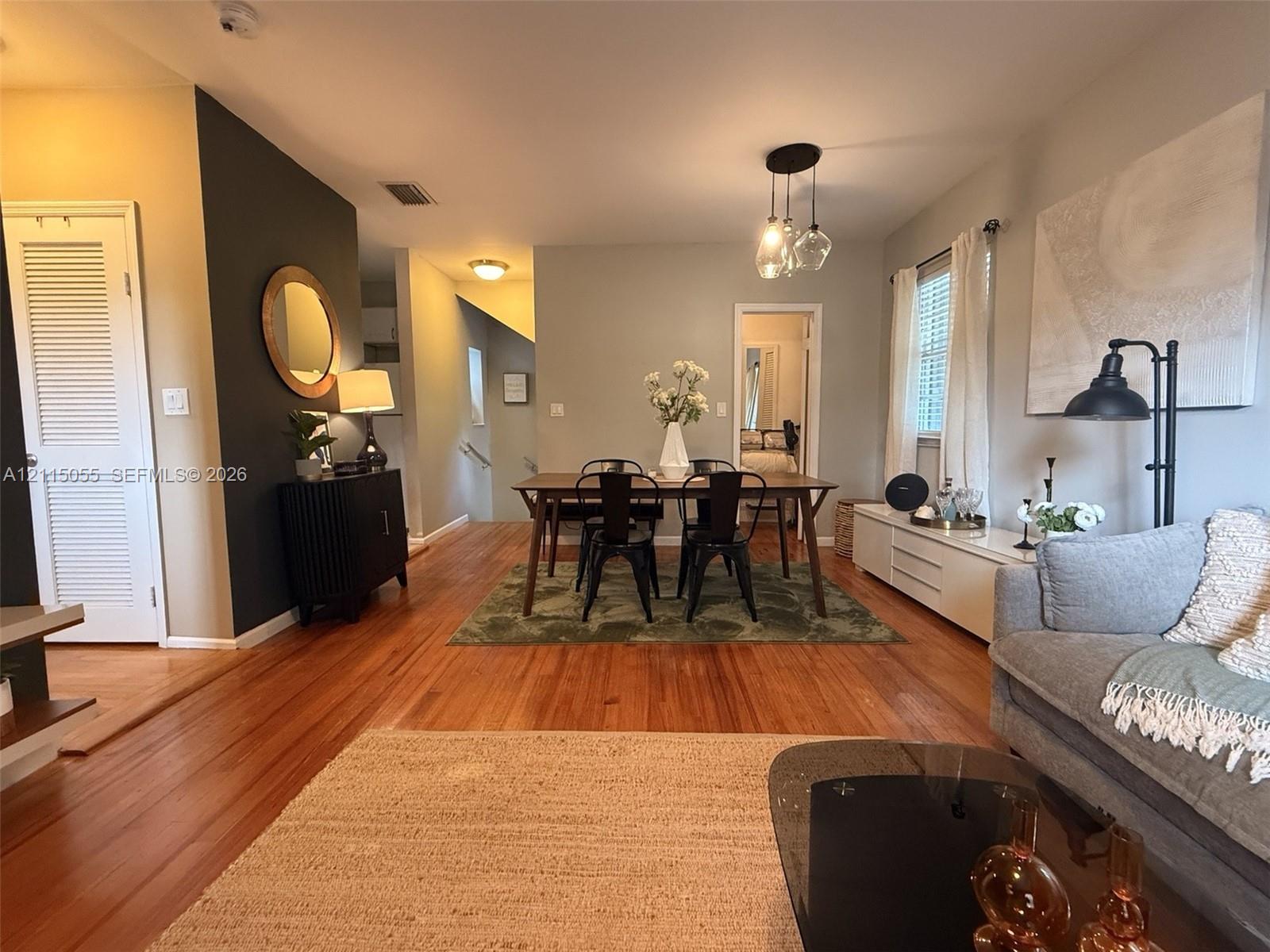 Open-concept living and dining area featuring wood-finish flooring, a dark charcoal accent wall, multi-pendant lighting, and louvered closet doors