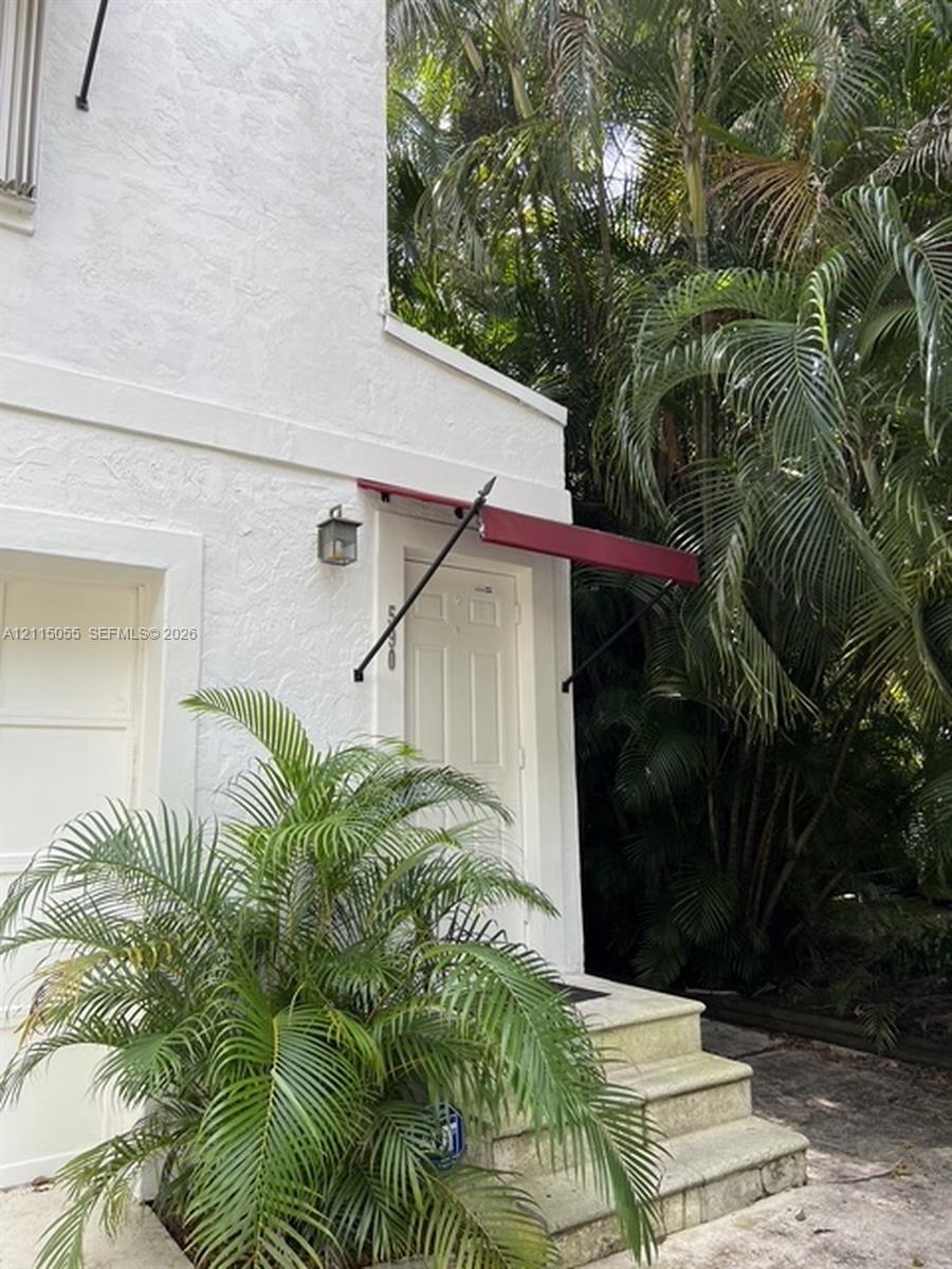 Stucco exterior featuring a six-panel entry door, metal awning, stone-finish steps, and a wall-mounted lantern fixture