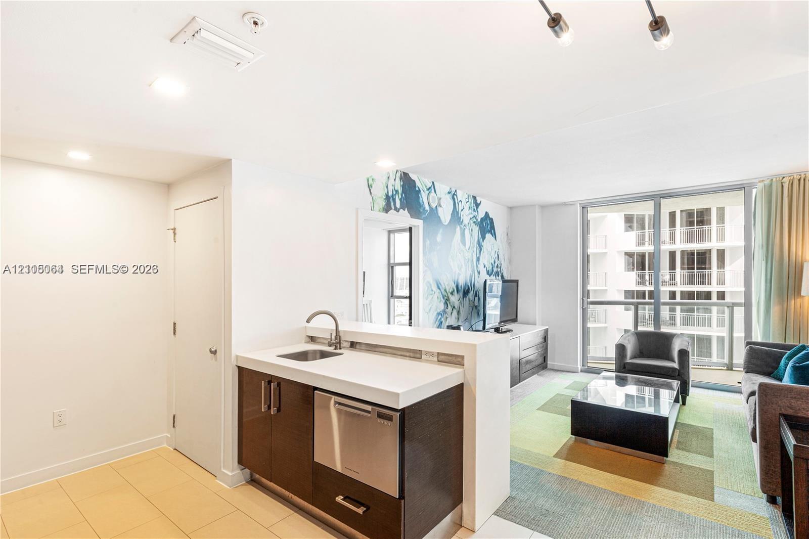 Kitchenette featuring dark cabinetry, solid-surface countertop, stainless steel undermount sink, and integrated stainless steel dishwasher, complemented by floor-to-ceiling glass panel windows