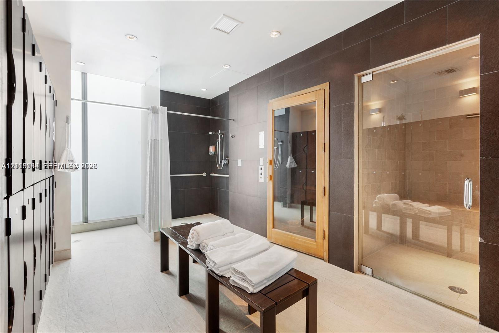 Spa area featuring a glass-enclosed steam room, a sauna with wood-framed glass door, floor-to-ceiling dark tile wall accents, built-in locker storage, and a shower area with wall-mounted plumbing fixtures