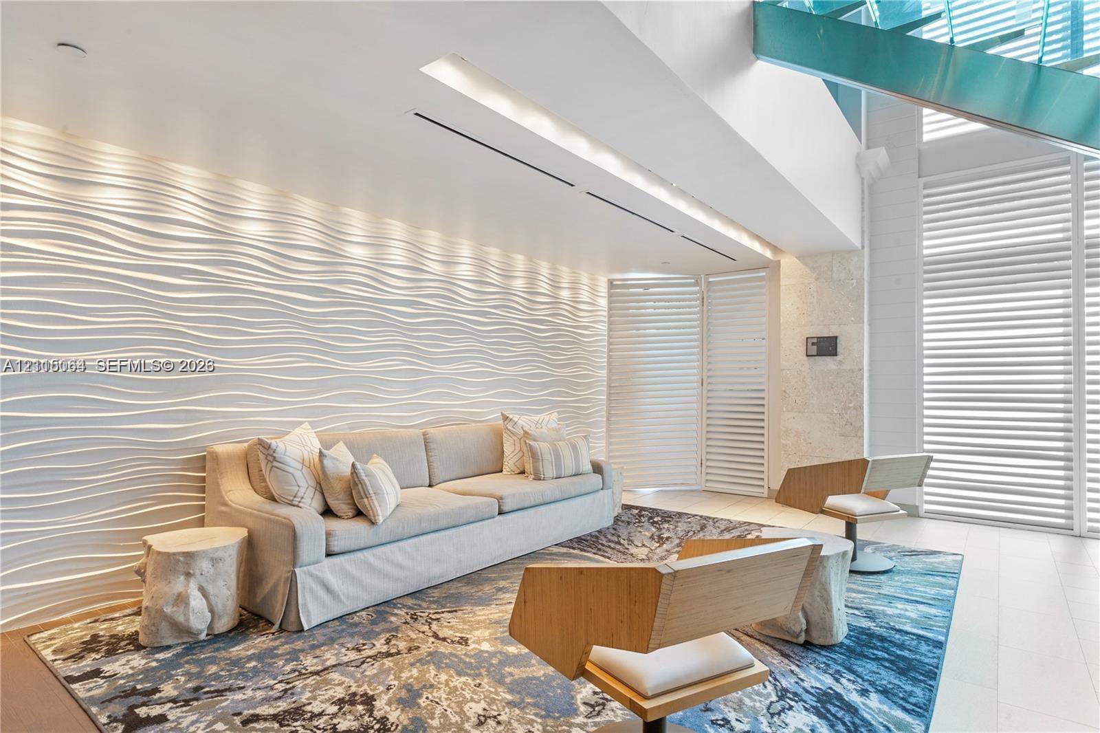 Textured wave-pattern accent wall, double-height ceilings, floor-to-ceiling louvered window treatments, glass-paneled staircase, and light-toned stone-finish flooring