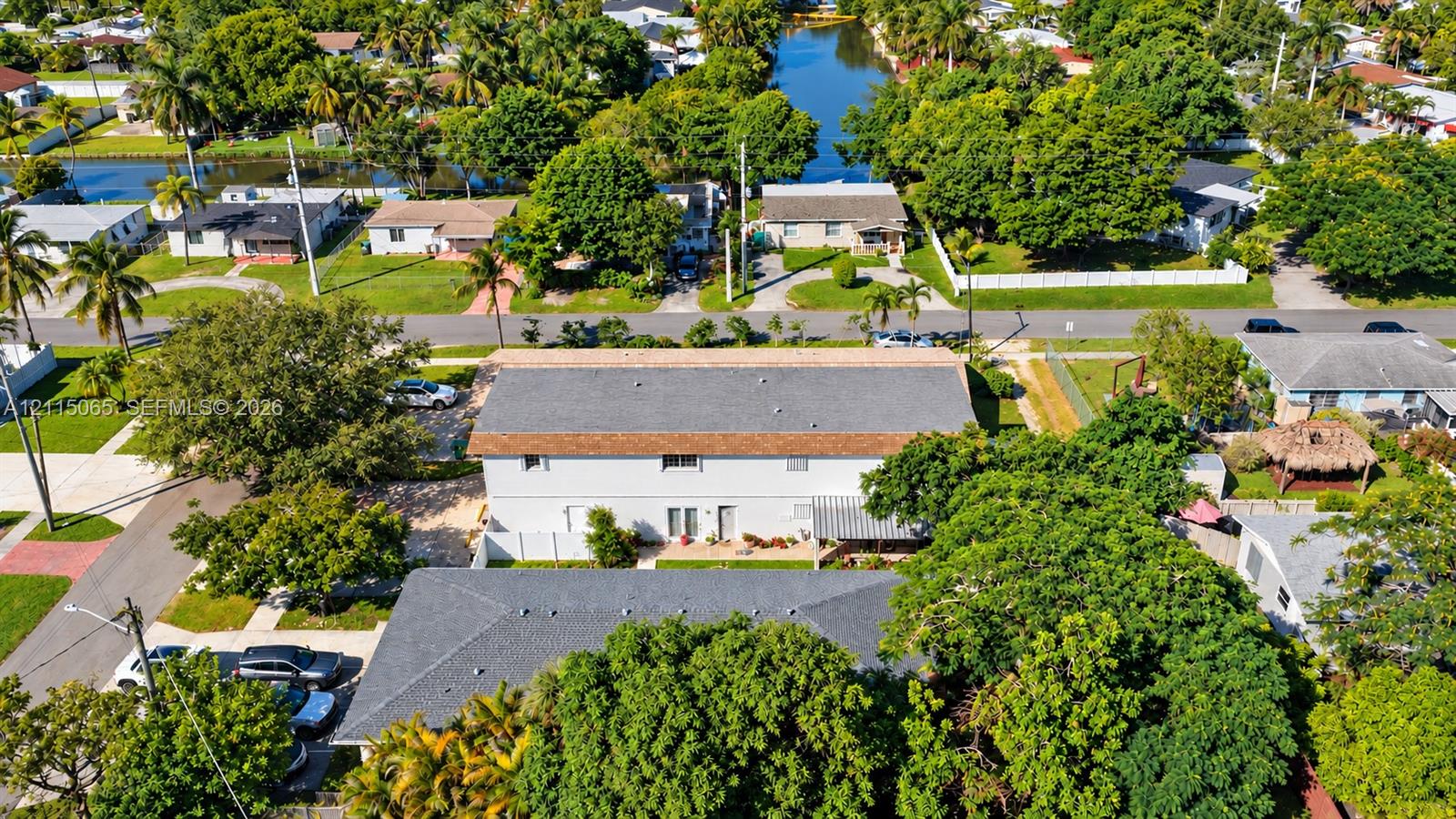 Aerial perspective of a multi-unit property featuring shingle roofing, paved off-street parking, a fenced perimeter, and mature canopy trees