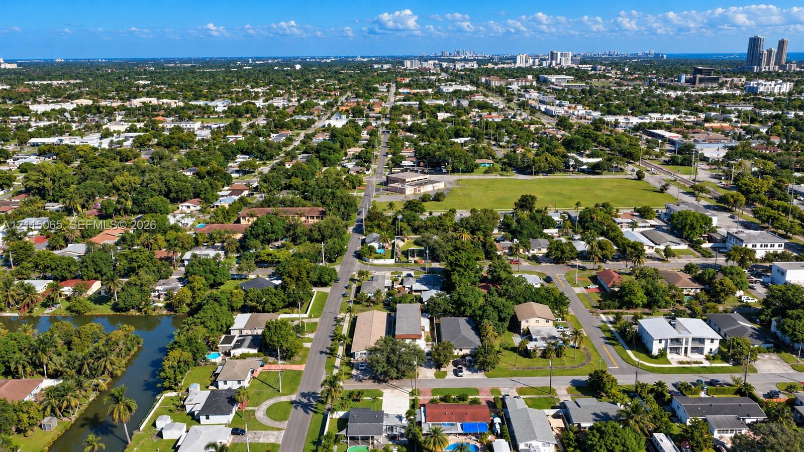 Aerial perspective of a residential neighborhood featuring mature tree canopies, direct water frontage, paved street grids, and proximity to urban skyline infrastructure
