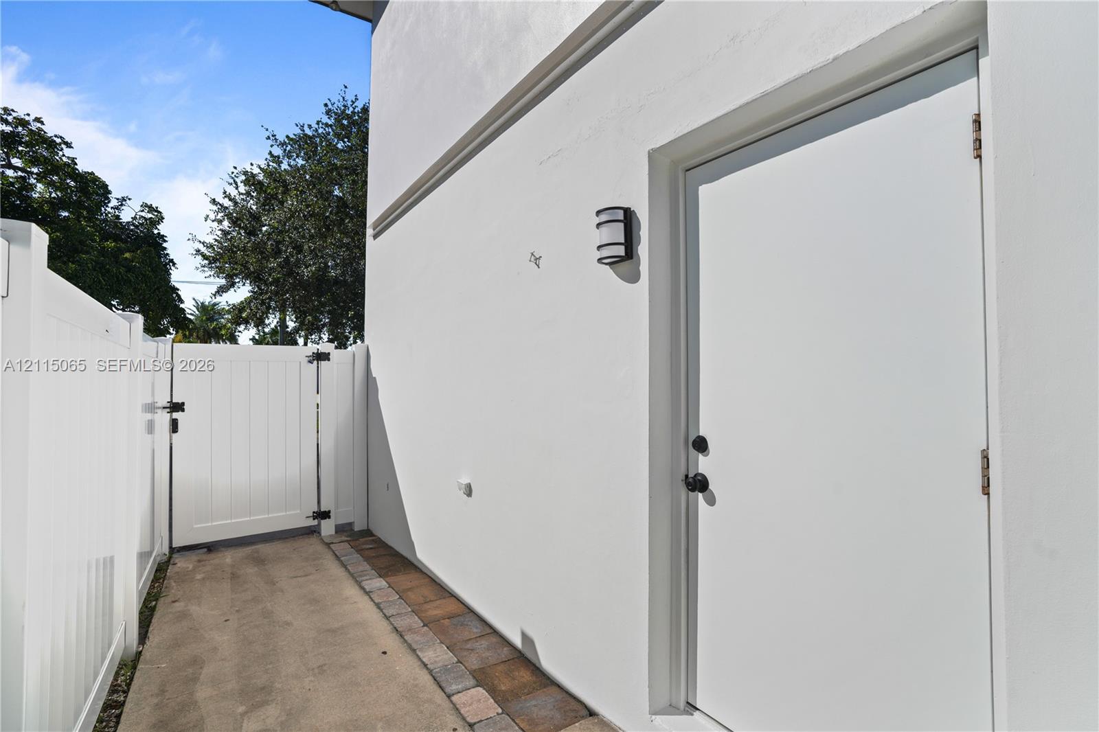 Exterior walkway featuring white stucco siding, a solid white side entry door, white vinyl privacy fencing, a matching white gate, and a paver stone border
