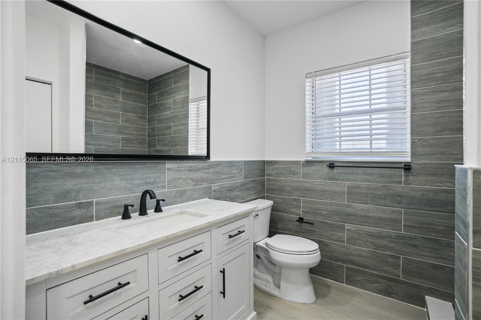 Bathroom vanity featuring white cabinetry, a stone countertop with an integrated rectangular sink, matte black hardware, and an expansive framed mirror, accented by horizontal grey stone-style wall tiling