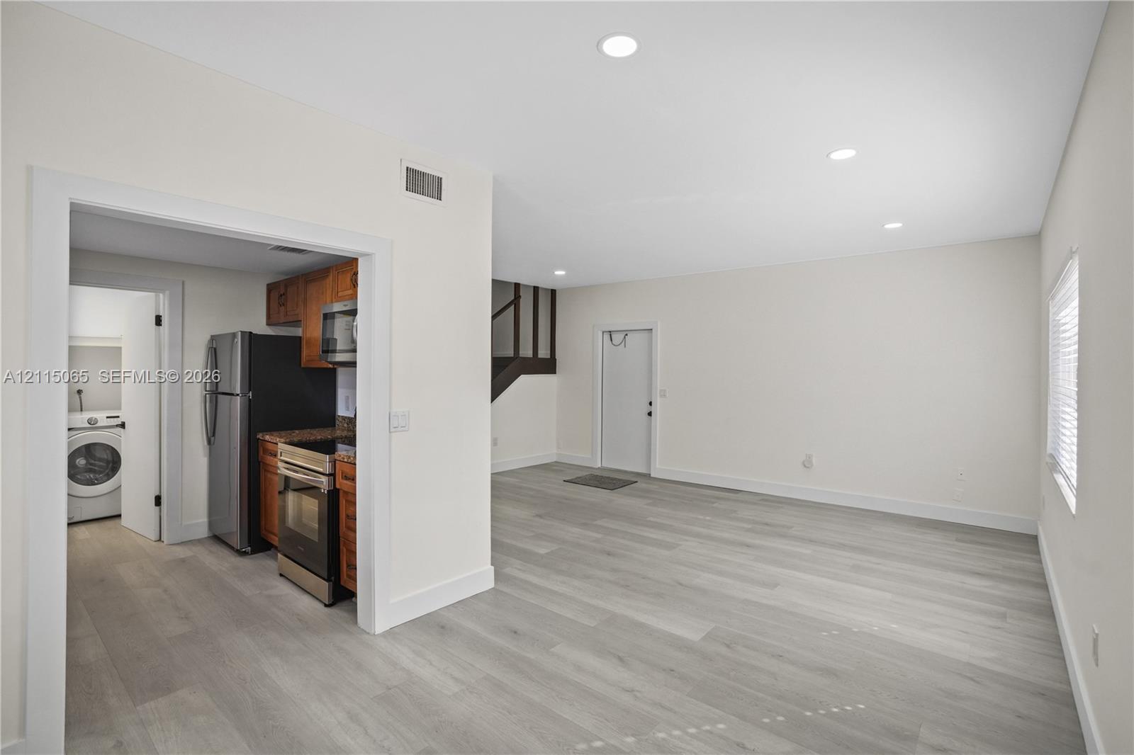 Open interior space featuring light wood-finish flooring, recessed lighting, and a kitchen equipped with stainless steel appliances, dark wood-tone cabinetry, and stone-surface countertops Open interior space featuring light wood-finish flooring, recessed lighting, and a kitchen equipped with stainless steel appliances, dark wood-tone cabinetry, and stone-surface countertops