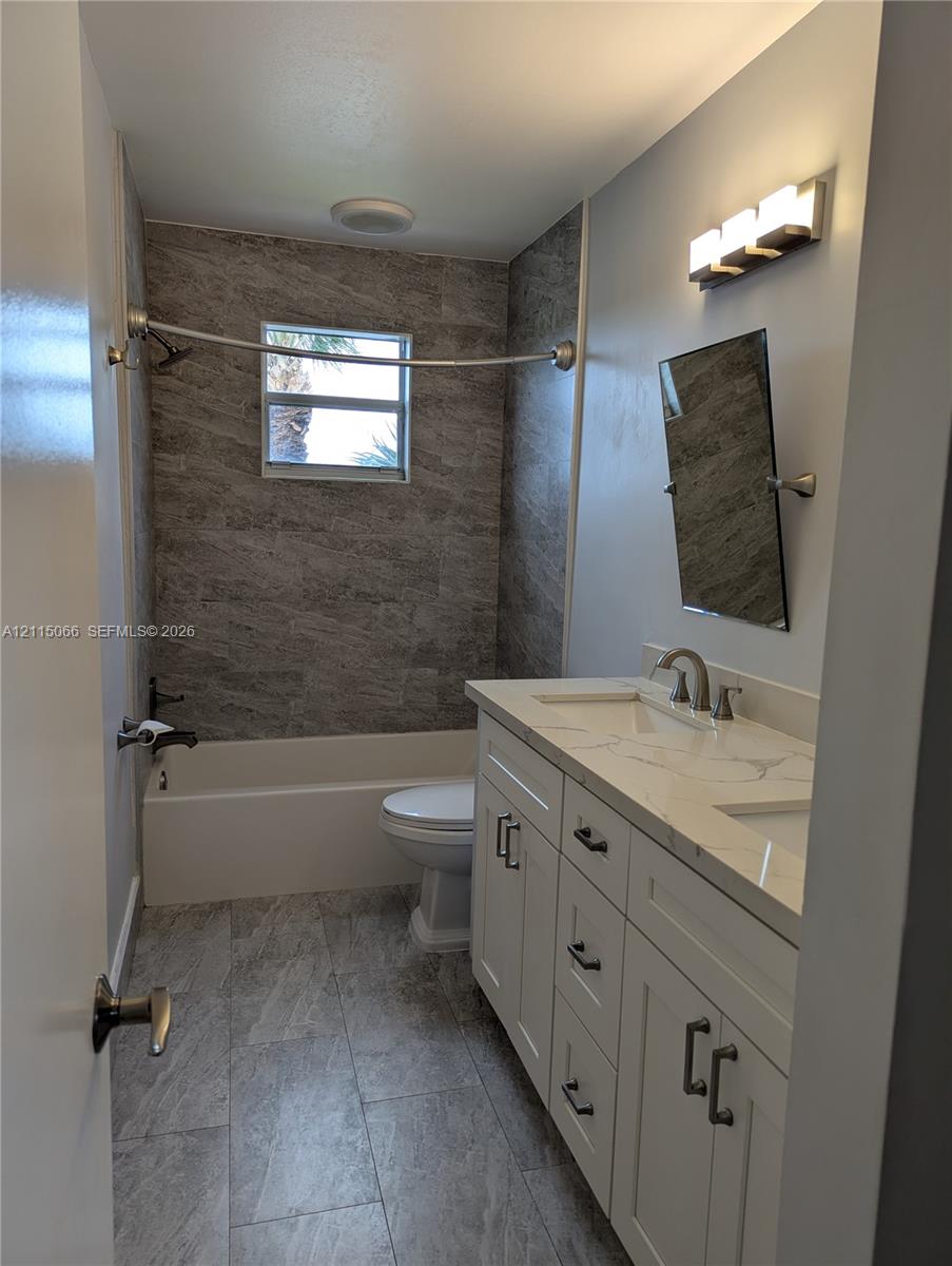 Bathroom featuring a dual-sink vanity with a stone-patterned countertop, grey stone-finish wall tile in the shower area, matching grey tile flooring, and a multi-bulb vanity light fixture