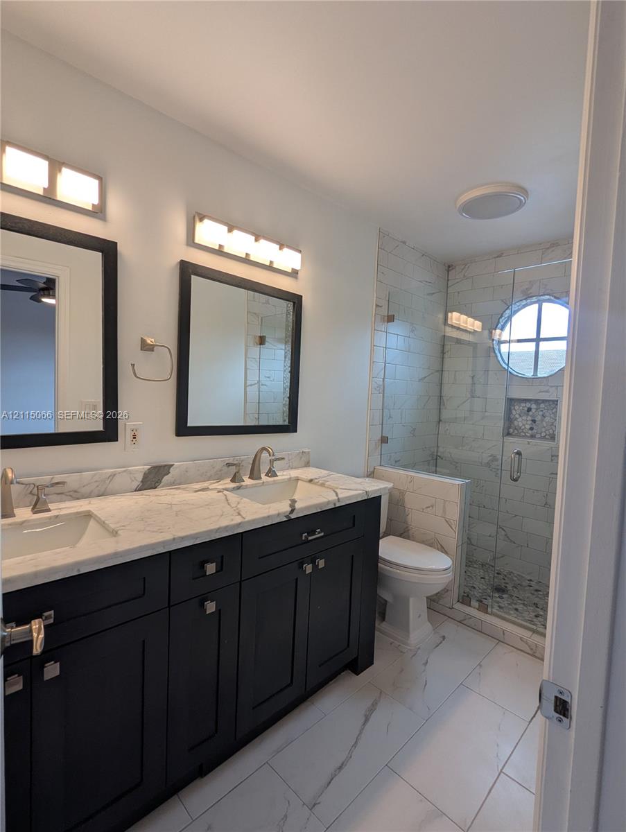 Double vanity with stone countertop and dual undermount sinks, custom dark cabinetry, large-format tile flooring, and walk-in glass-enclosed shower featuring a porthole window and decorative tile inlay