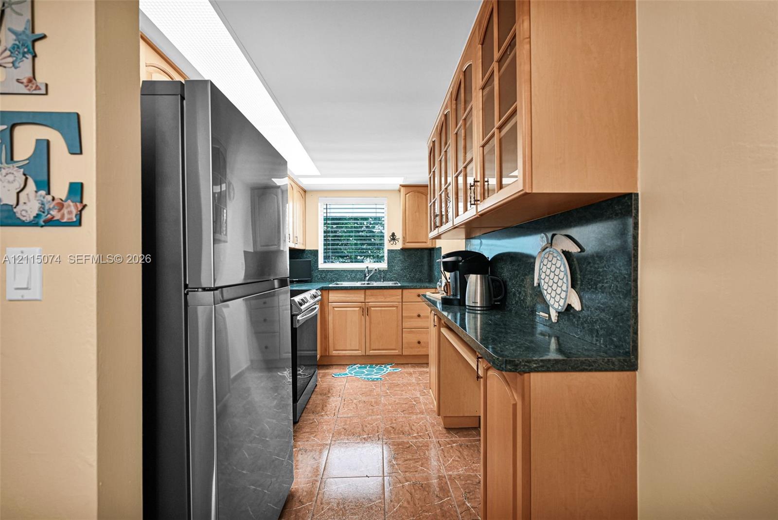 Kitchen featuring natural wood cabinetry with glass-front upper displays, dark countertops, tiled flooring, stainless steel refrigerator, and recessed lighting