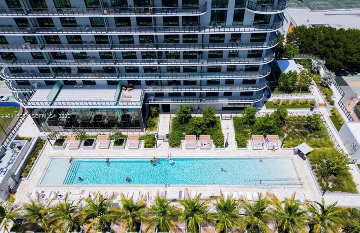 High-rise residential exterior featuring a multi-story glass facade with private balconies, a rectangular swimming pool, integrated landscaping with mature palm trees, and paved outdoor lounging areas High-rise residential exterior featuring a multi-story glass facade with private balconies, a rectangular swimming pool, integrated landscaping with mature palm trees, and paved outdoor lounging areas