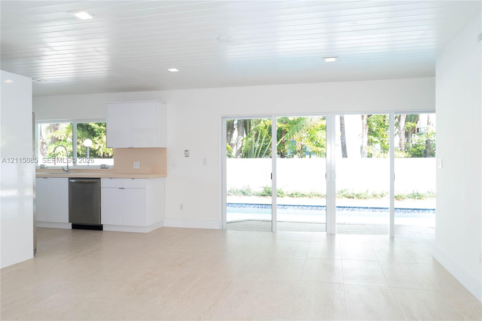Kitchen featuring white cabinetry, light-toned countertops, and stainless steel dishwasher, with light-toned porcelain tile flooring and sliding glass doors opening to a swimming pool