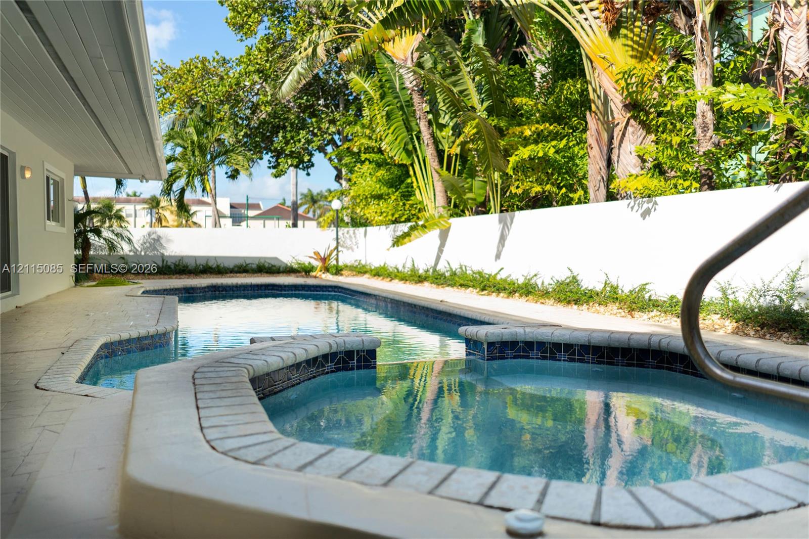 In-ground swimming pool and attached spa featuring decorative tile accents, light-toned stone coping, surrounding hardscape patio, and a privacy wall