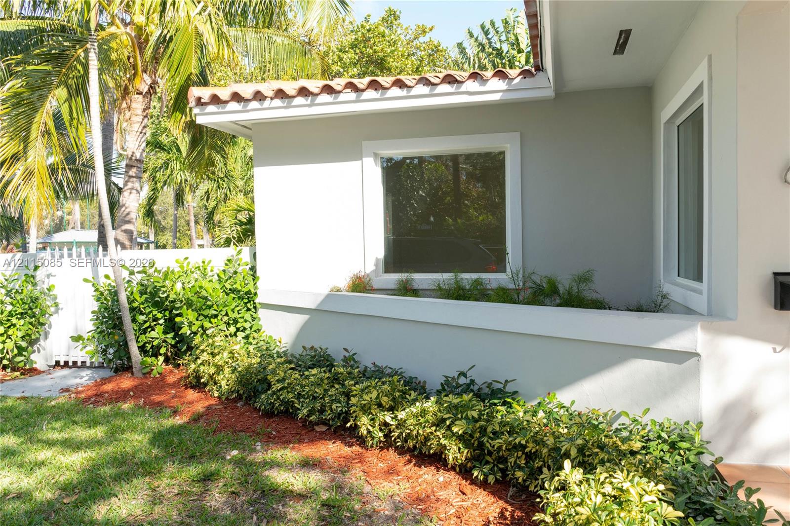 Exterior featuring light-toned stucco siding, a terracotta tile roofline, crisp white window trim, and a manicured foundation garden with mulch beds