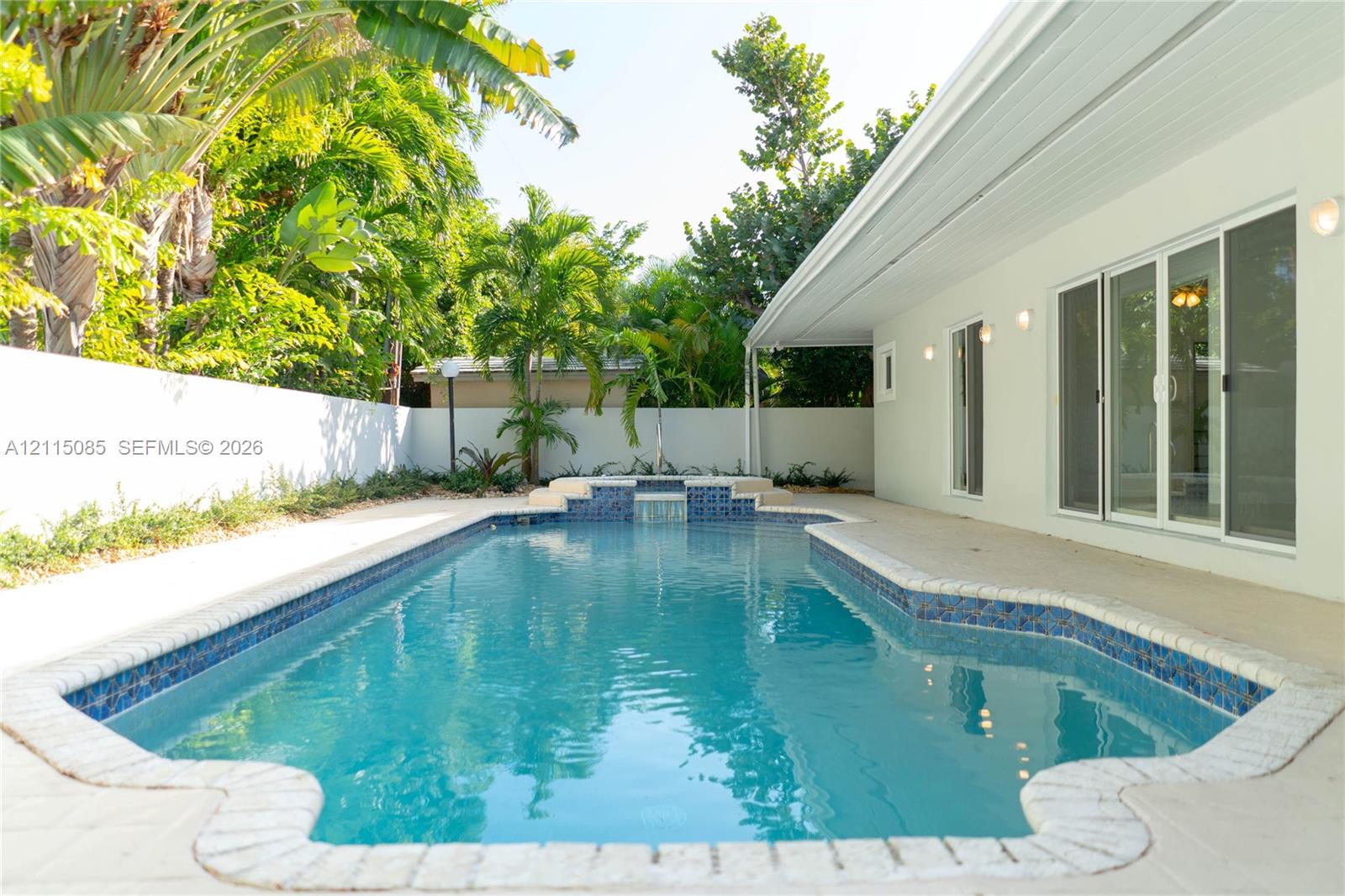 In-ground swimming pool featuring a tiled waterfall element, stone-paver decking, and a perimeter privacy wall