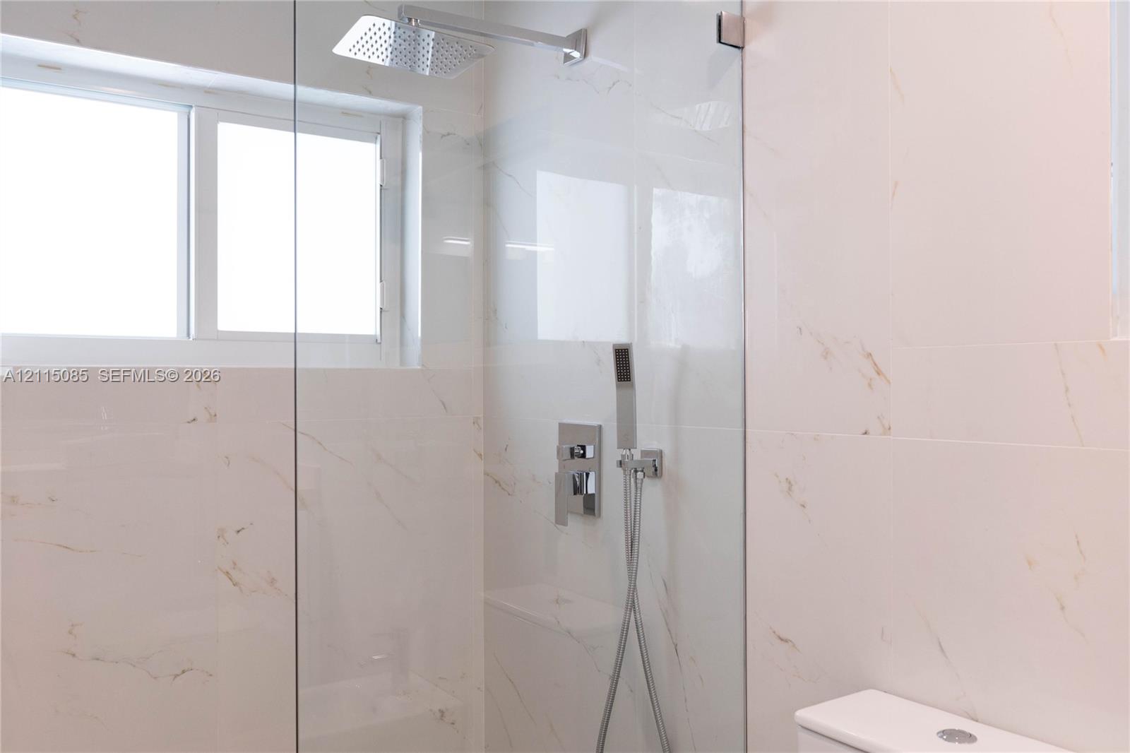 Walk-in shower featuring large-format stone-finish wall tile, frameless glass enclosure, rainfall showerhead, handheld shower wand, and integrated chrome fixtures