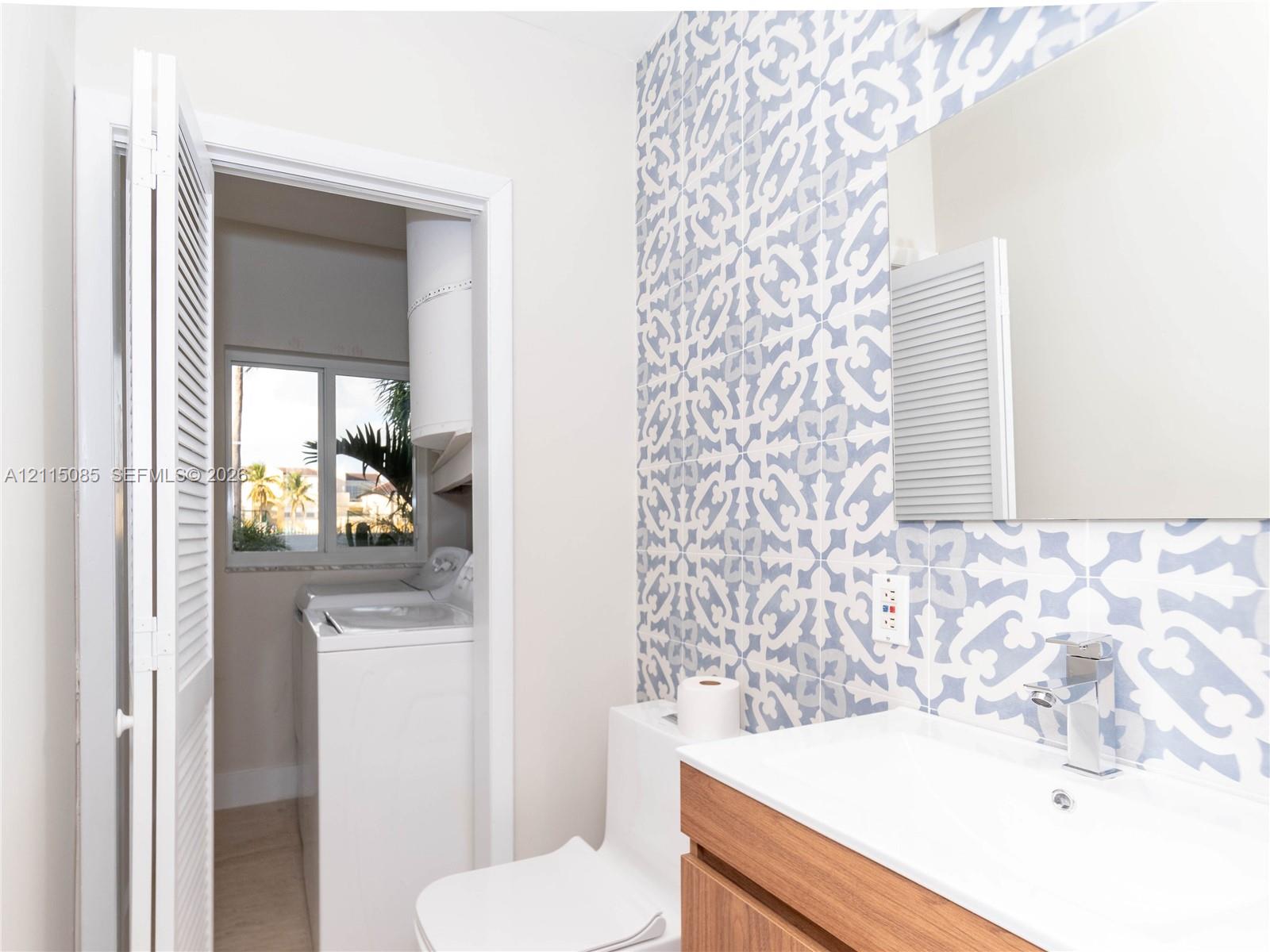 Bathroom featuring a wood-finish vanity with a rectangular sink, wall-mounted frameless mirror, patterned tile accent wall, and a louvered closet door concealing laundry hookups