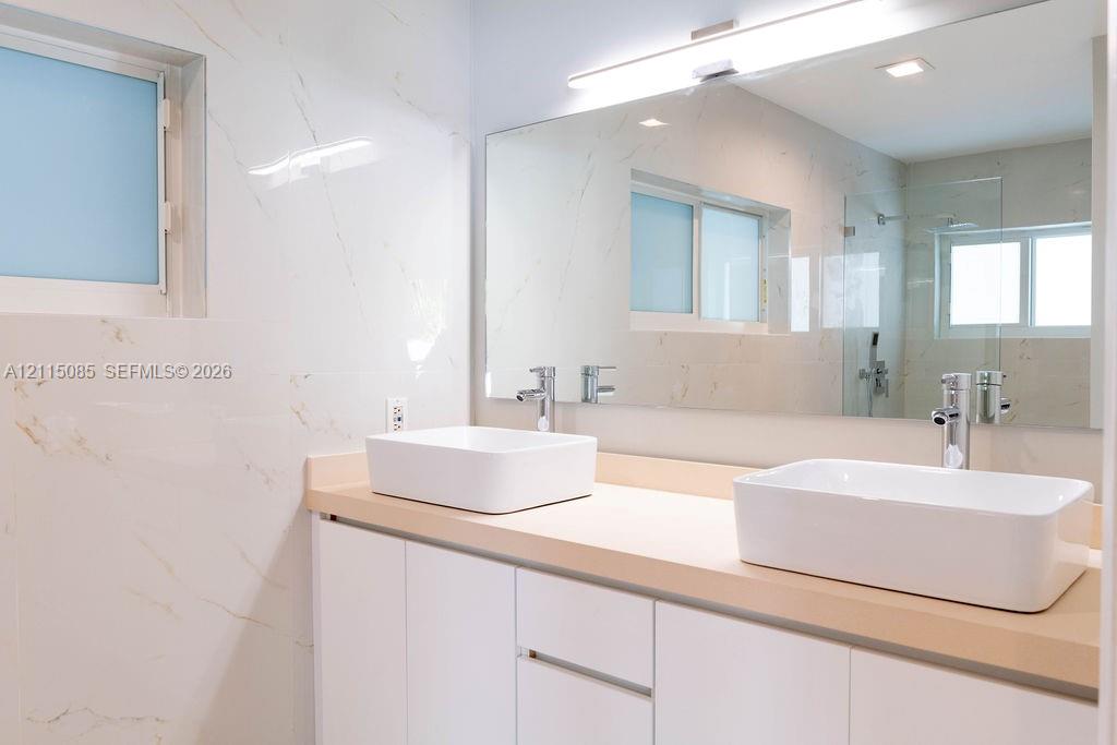 Double vanity featuring white vessel sinks, polished chrome faucets, a solid-surface countertop, white cabinetry, and wall-to-wall mirror with integrated linear lighting, all accented by large-format, veined tile wall finishes