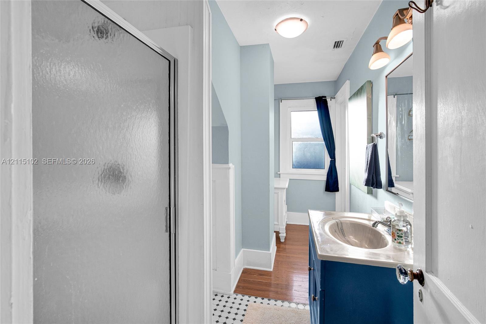 Bathroom featuring a vanity with an integrated basin, a walk-in shower with a framed glass door, wood-finish flooring, a mosaic tile floor section, and wall-mounted light fixtures