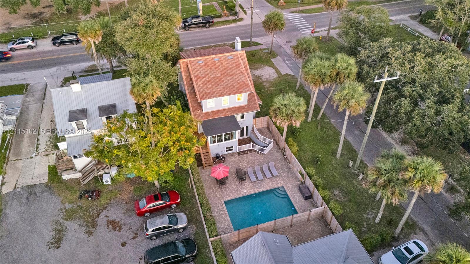 Aerial view featuring a multi-story residence with a red-toned roof, an in-ground swimming pool, a stone-paver patio, and mature palm trees